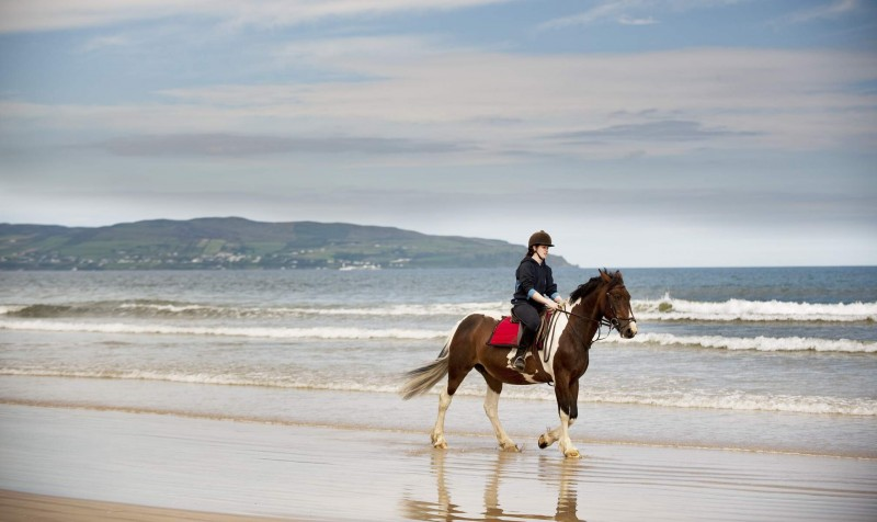 A horse and rider making their way along Benone beach, with waves crashing along the shoreline. Credit: Tourism Ireland by Gardiner Mitchell.