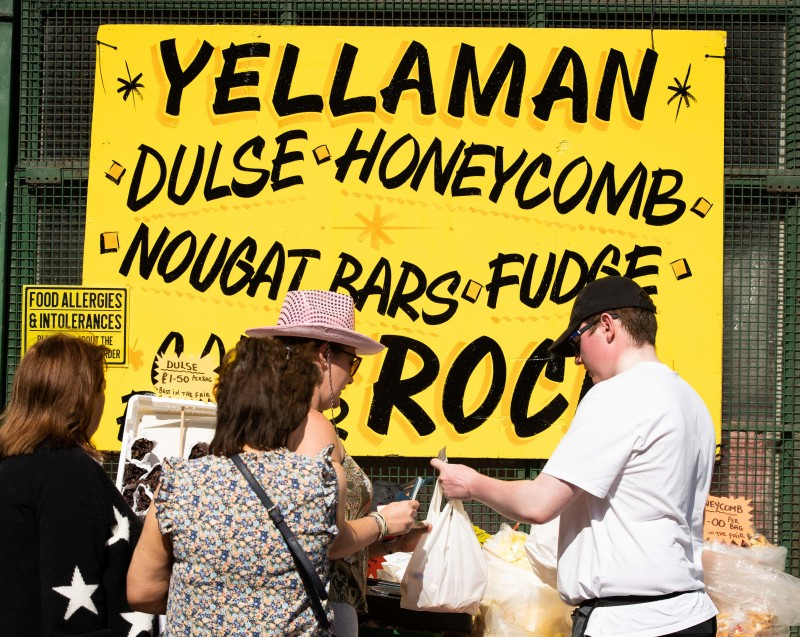 Visitors purchasing sweet treats including Yellowman and Dulse. Sign in the background reads “Yellaman, Dulse, Honeycomb, Nougat Bars, Fudge and Rock”.