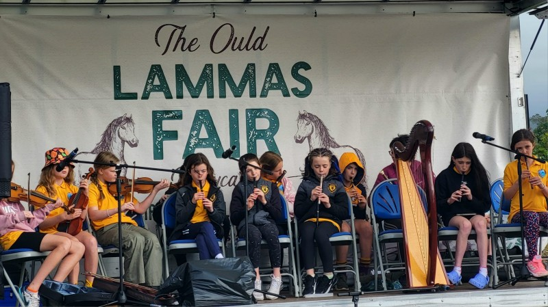 A group of young Irish Trad musicians on stage at The Seafront, with a sign behind them reading “Ould Lammas Fair, Ballycastle”.