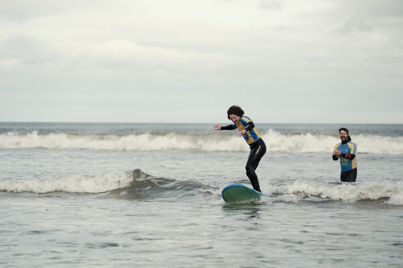 A man standing on a surfboard in the waves, with an instructor encouraging them from behind. Credit: Tourism Northern Ireland.